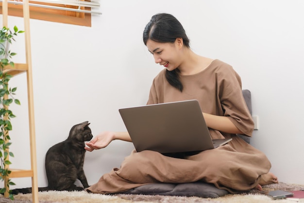 Pet lover concept Young Asian woman using laptop and playing to stroking with cat in living room