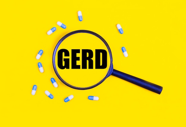 Pharmaceutical tablets and capsules near a magnifying glass with the text GERD on a yellow background View from above