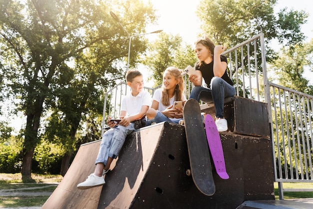 Phone addicted sports children with skateboard and penny boards smiling and looking at smartphones on sports ramp on playground Kids addiction of phones