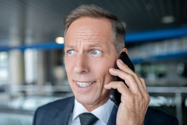 Phone conversation. Business attractive adult man in formal suit and tie talking on smartphone in an airport building during day