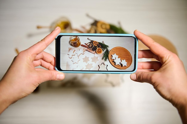 Phone in real time. Close-up. Hands holding phone and taking pictures of gingerbreads in the shape of stars laid out on wooden board and on baking paper next to dried orange slices, pine cones, twigs