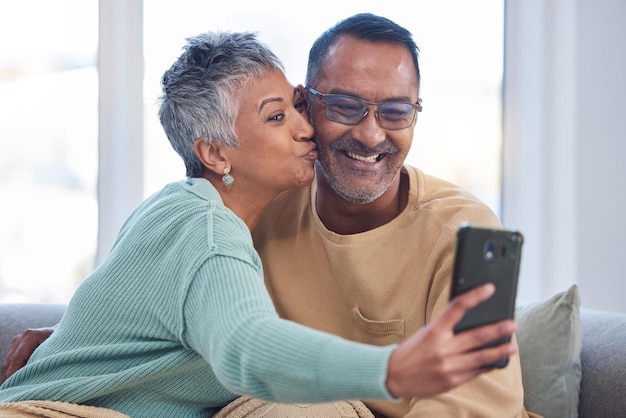 Phone selfie and kiss with a senior couple posing for a picture in the living room of their home together Smartphone kissing and social media with a mature man and woman taking a photograph