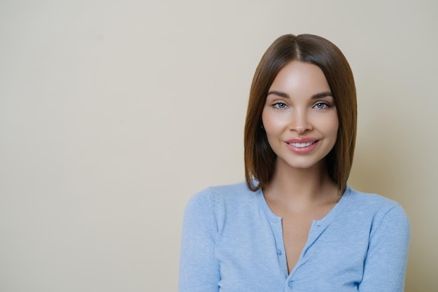 Photo of adorable brunette woman has natural makeup and healthy perfect skin smiles tenderly shows white teeth spends free time on caring about herself wears casual blue jumper poses indoor