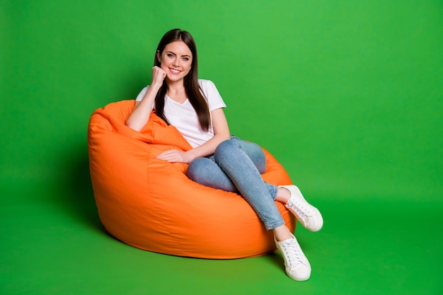 Photo of adorable cute brunette hair young woman fist cheek dress wear white t-shirt jeans footwear sit on pouf isolated on green background