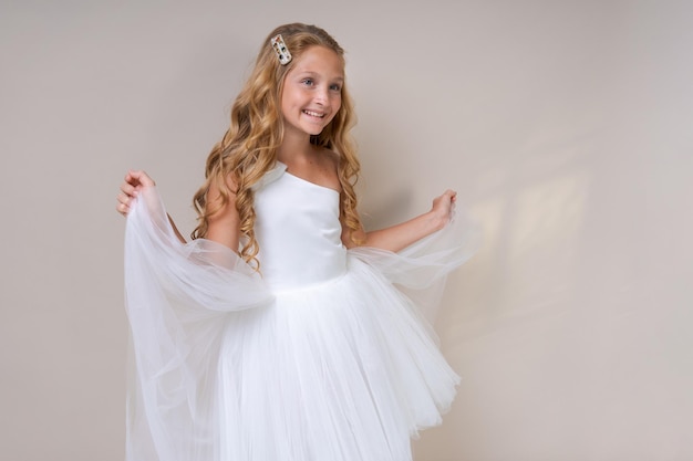 Photo an adorable cute schoolgirl angel in white dress, on a light background. Caucasian beautiful girl posing happy in the studio. Angel child