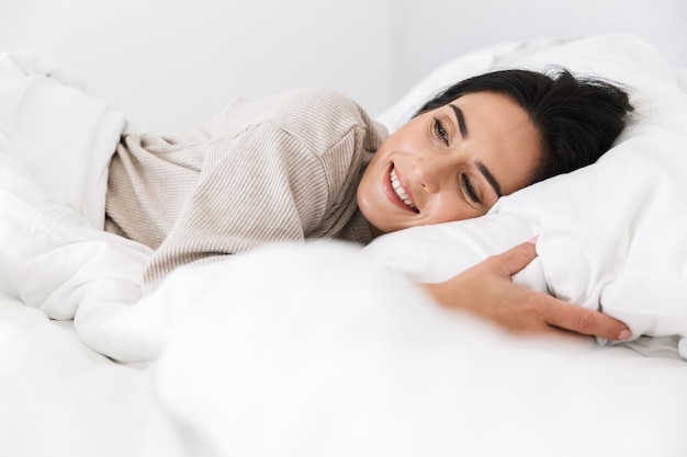 Photo of adorable woman 30s smiling, while lying in bed with white linen at home