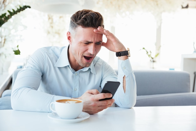 Photo of adult man being excited and puzzled while reading text message or news on black mobile phone, when sitting alone in city cafe with cup of coffee