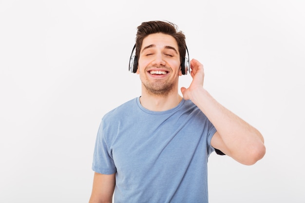 Photo of adult smiling guy having short dark hair enjoying music via earphones with closed eyes, isolated over white wall