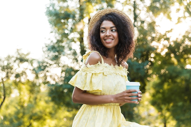 Photo of african american woman 20s wearing straw hat, walking outdoor and holding takeaway coffee