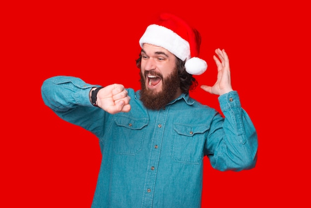 Photo of amazed bearded man with santa claus hat looking at watch