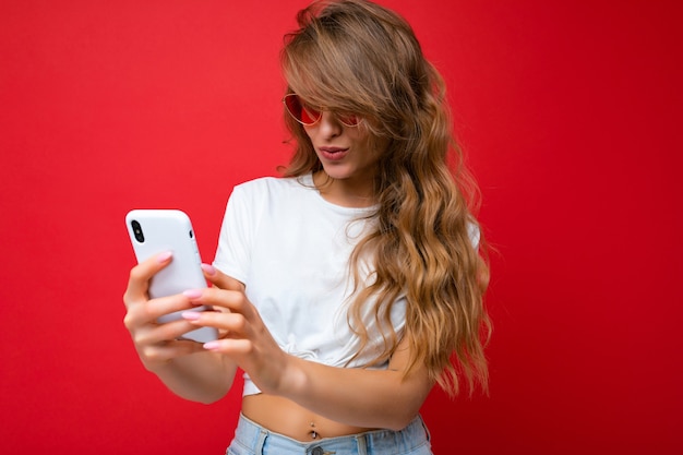 Photo of amazed beautiful young woman holding mobile phone wearing sunglasses everyday stylish outfit isolated over wall background looking at device screen and communicating