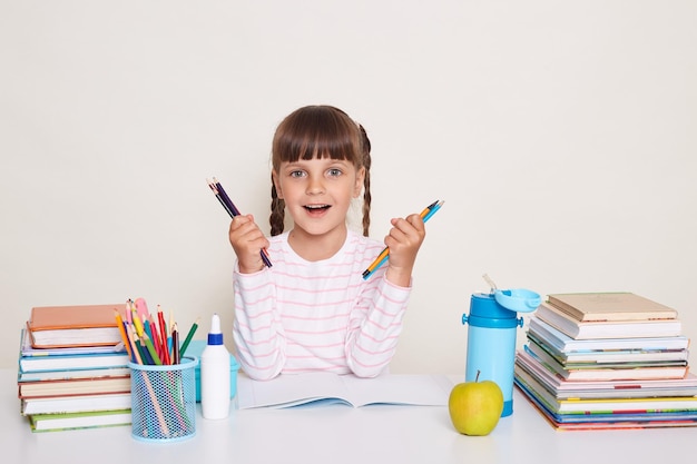 Photo of amazed little schoolgirl with pigtails wearing striped shirt sitting at the desk surrounded with school supplies holding pens or pencils looking at camera with optimistic happy expression