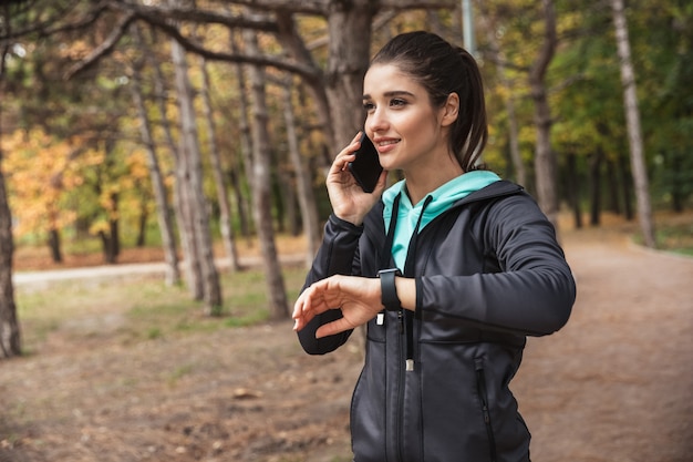 Photo of amazing young pretty fitness woman outdoors in the park looking at watch clock talking by mobile phone.