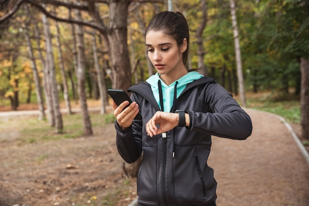 Photo of amazing young pretty fitness woman outdoors in the park looking at watch clock using mobile phone.