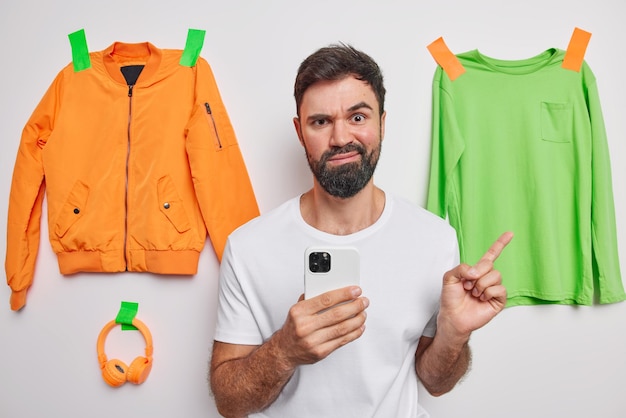 Photo of angry dissatisfied bearded young man holds mobile phone points index finger at items of clothes sells uncessary clothing in internet wears casual white t shirt recommends something.