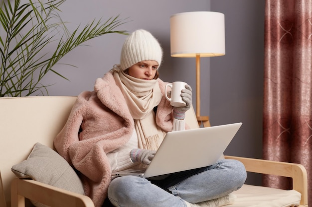 Photo of angry frozen woman wearing cap gloves scarf and coat sitting in home interior with cup of hot tea trying to warm up working on laptop looking at display with anger and hate