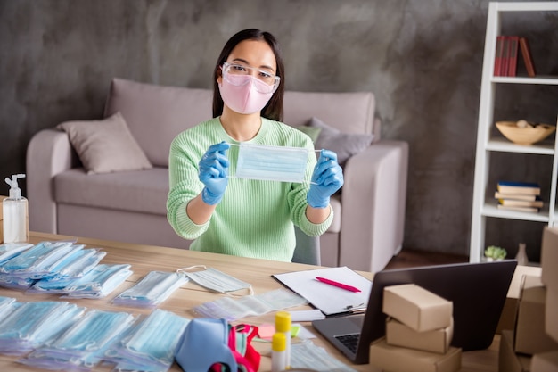 Photo of asian business lady blogger organize orders facial flu masks showing ready product web camera process preparing packs delivery package safety home office quarantine indoors