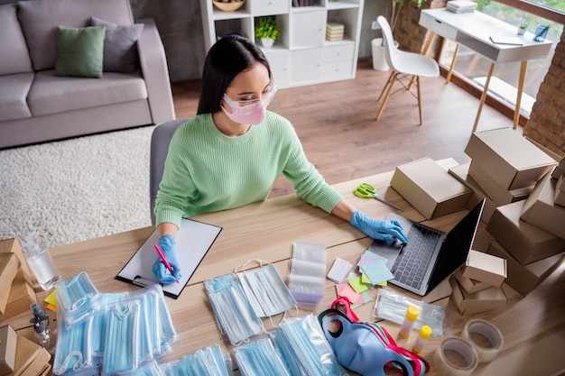 Photo of asian business lady organize read write notebook orders manager facial flu masks product internet service preparing delivery package safety home office quarantine indoors