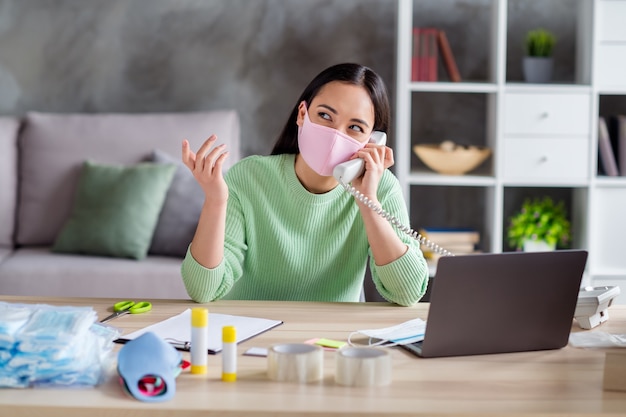 Photo of asian busy business lady organizing facial flu medical masks packs for delivering boxes speaking pick up landline customer write clipboard order details home office indoors