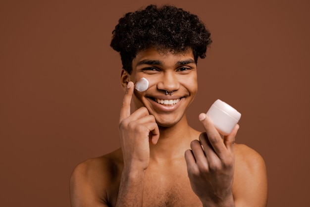 Photo of attractive black man with piercing smiles and put nutrition cream on face. Naked torso, isolated brown color background.