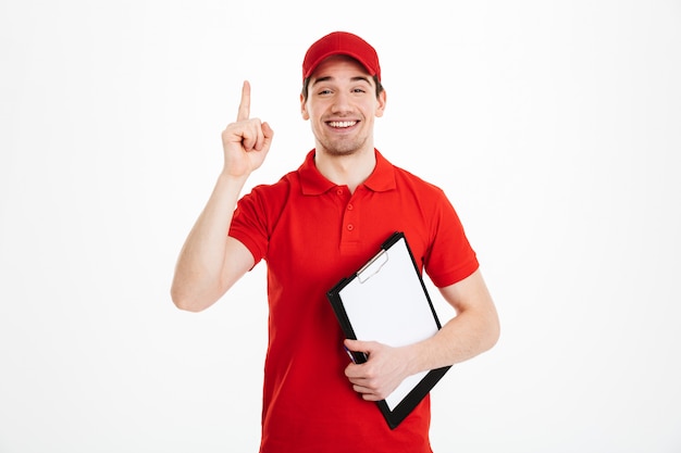 Photo of attractive delivery worker in red t-shirt and cap smiling and pointing finger up on copyspace text or product while holding clipboard, isolated over white space