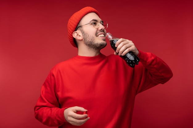 Photo of attractive man with beard in glasses and red clothing. Happy man holds drink, isolated over red background.