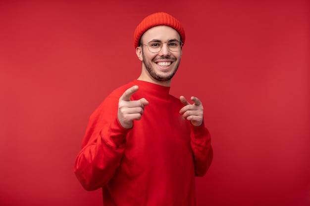 Photo of attractive man with beard in glasses and red clothing. Male snaps fingers and smiles, isolated over red background.