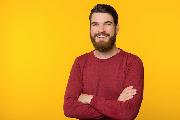 Photo of bearded guy, standing with crossed arms, looking and smiling at camera  over yellow isolated background