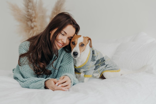 Photo of beautiful brunette woman in knitted sweater lies together wih dog on soft bed enjoys spending time with favorite pet cares about animals stay at home during coronavirus quarantine