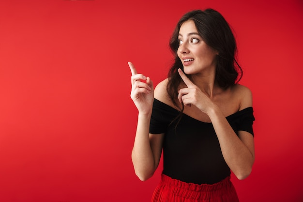 Photo of a beautiful cute young woman standing isolated over red wall pointing.