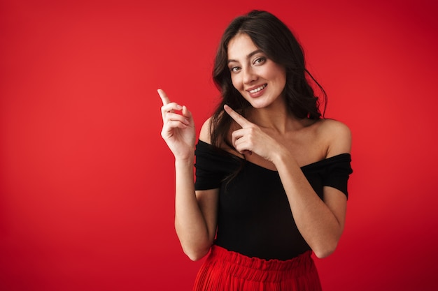 Photo of a beautiful cute young woman standing isolated over red wall pointing.