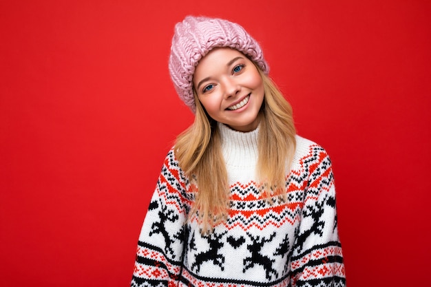 Photo of beautiful happy smiling young blonde woman isolated over red background wall wearing winter sweater and trendy pink hat looking at camera.