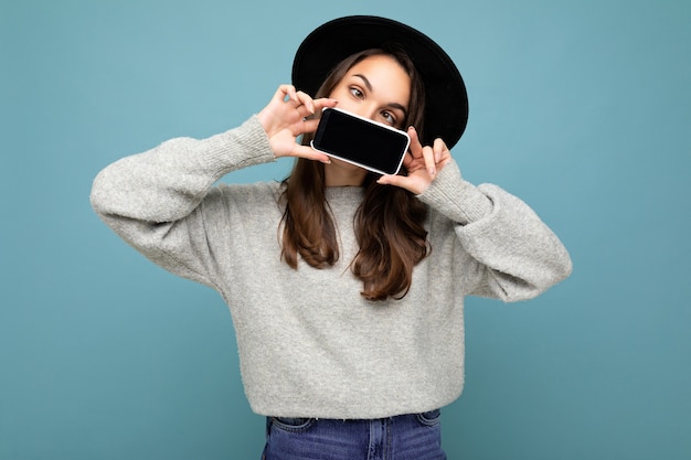 Photo of Beautiful positive young female person wearing black hat and grey sweater holding mobilephone showing smartphone isolated on background looking at camera