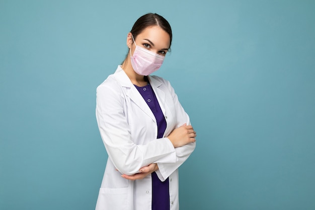 Photo of beautiful young woman doctor in white coat and medical mask standing isolated over blue background. Coronavirus concept