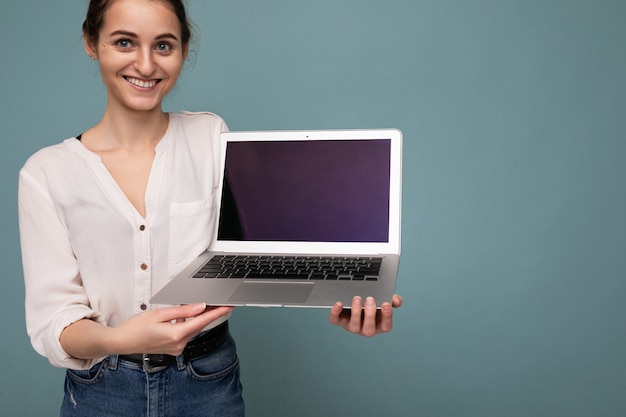 Photo of beautiful young woman holding computer laptop looking at camera isolated over colourful background.