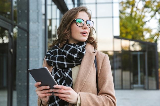 Photo of a beautiful young woman outdoors walking by street using tablet computer.