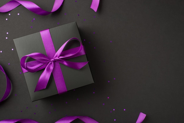 Above photo of black giftbox with purple ribbon wrapped as bow and confetti isolated on the black background