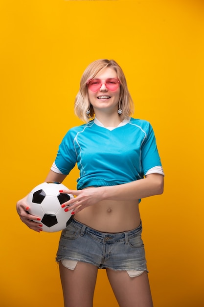 Photo photo of blonde in pink glasses with soccer ball on orange backgroundin studio