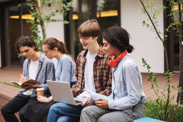 Photo of boy and girl sitting on bench and using laptop while spending time in courtyard of university