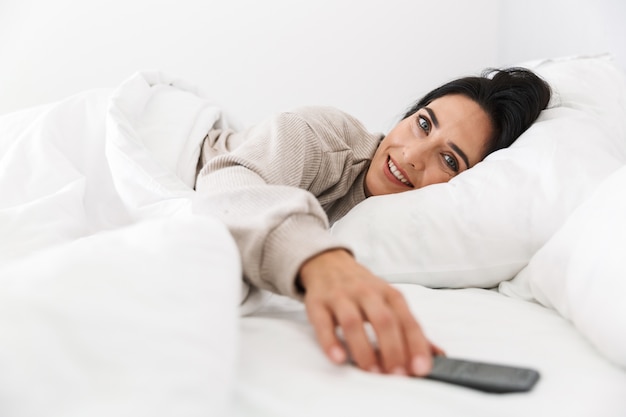 Photo of brunette woman 30s using smartphone, while lying in bed with white linen at home