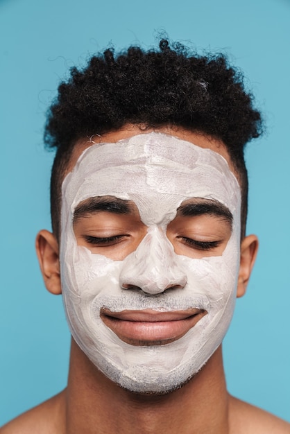 Photo of calm african american man in facial cosmetic mask smiling with eyes closed isolated on blue