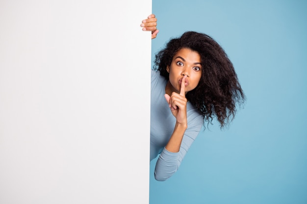 Photo of casual curly wavy beautiful brown haired girl showing you shh sign with forefinger touching her lips looking out of white banner isolated blue pastel color background