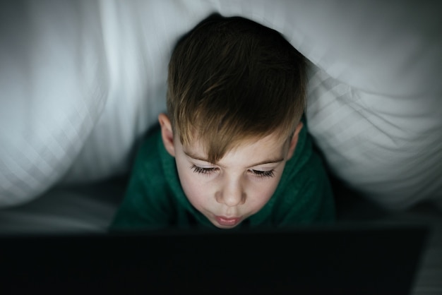 Photo of caucasian happy kid hiding under blanket playing computer