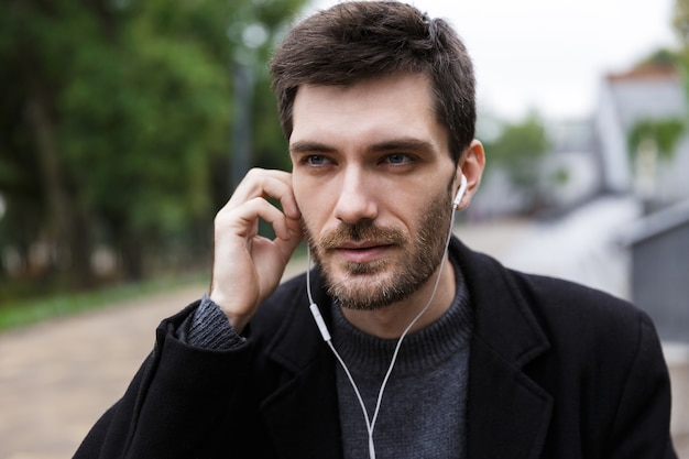 Photo of caucasian man 20s wearing earphones using mobile phone, while walking outdoor