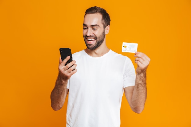 Photo of caucasian man 30s in casual wear holding smartphone and credit card, isolated