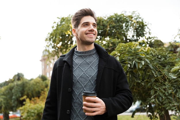 Photo of caucasian man 30s wearing jacket holding paper cup of takeaway coffee while walking through green park