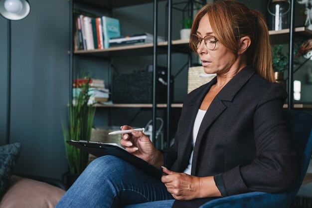 Photo of caucasian smart psychologist woman wearing eyeglasses sitting on armchair and holding clipboard in cabinet