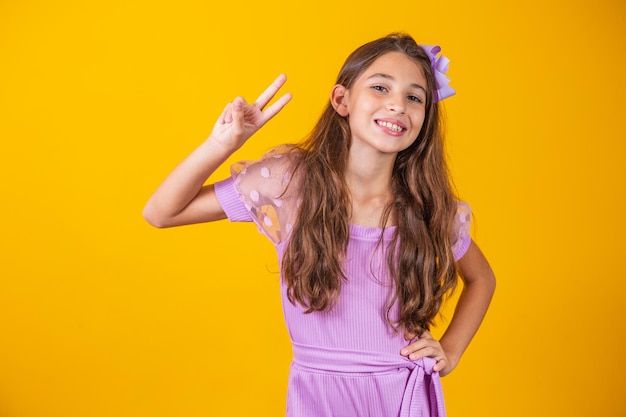 Photo of a charming and delicate preteen girl making a V sign smiling isolated on yellow background.