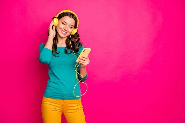 Photo of cheerful nice pretty cute charming girlfriend listening to music in her headphones holding telephone with hands isolated pink vibrant color background