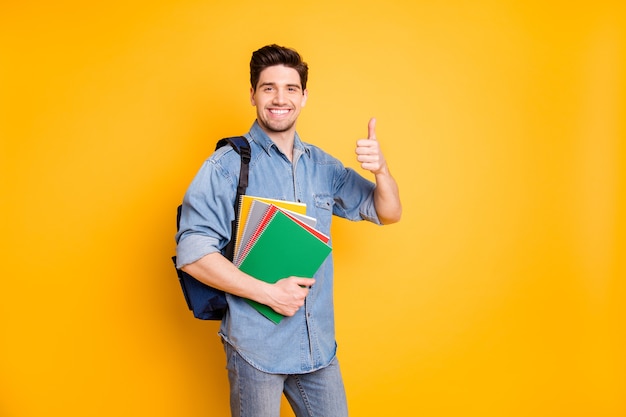 Photo of cheerful positive handsome man showing you thumb up holding copybooks with backpack behind smiling toothily isolated vivid color wall